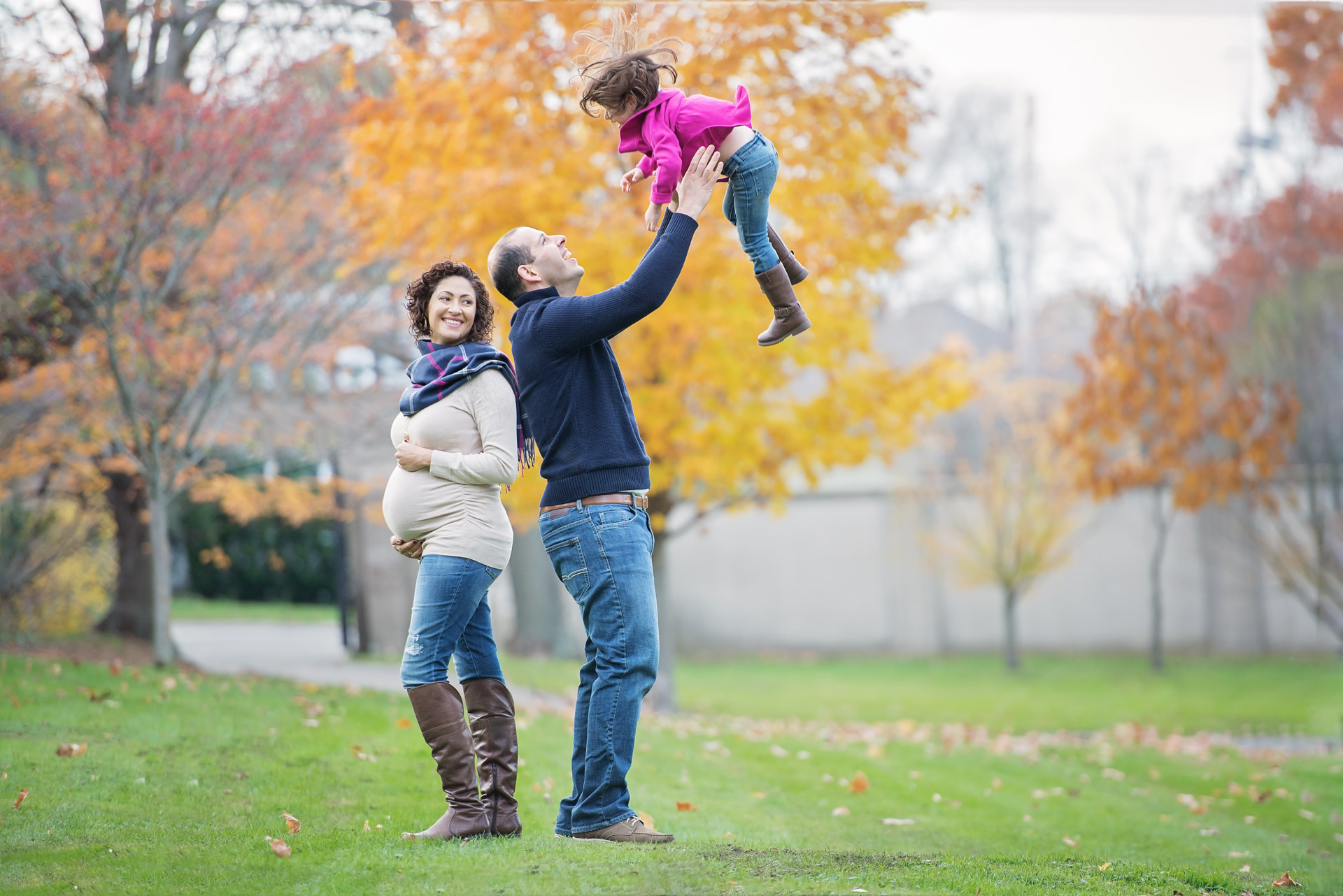 Father lifting his young daughter overhead while her expecting mother watches, under orange maples at Larz Anderson Park in Brookline MA