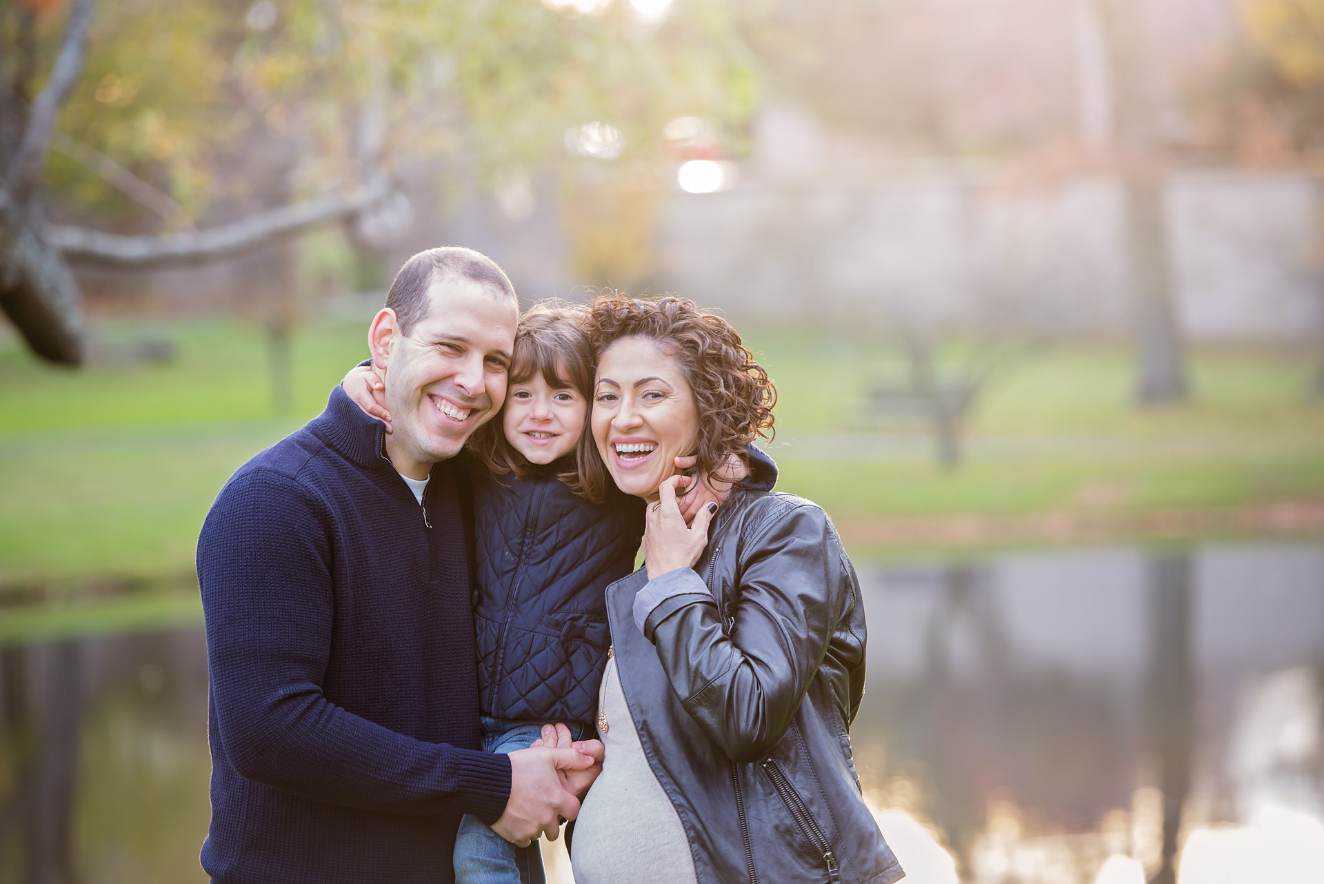 Expecting mother, her husband and their young daughter laughing together at the edge of the lagoon in low autumn light at Larz Anderson Park in Brookline MA