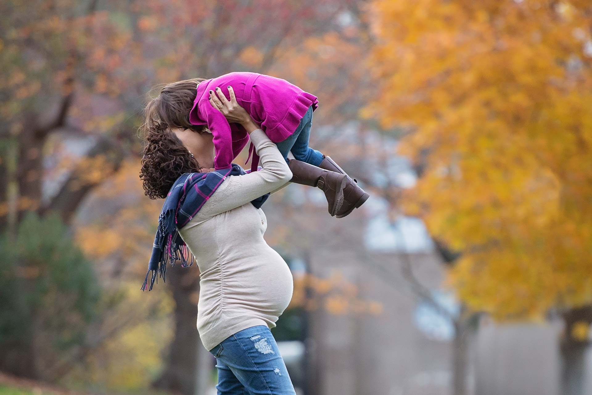 Expecting mother lifting her young daughter into the air beneath a gold maple at Larz Anderson Park in Brookline Massachusetts
