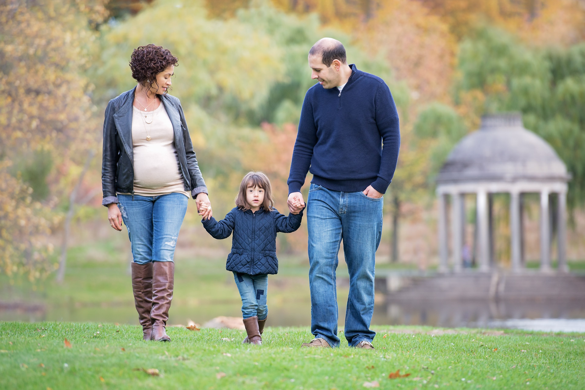 Expecting mother and her husband walking across the lawn holding their young daughter&rsquo;s hands with the Temple of Love rotunda behind them at Larz Anderson Park in Brookline MA