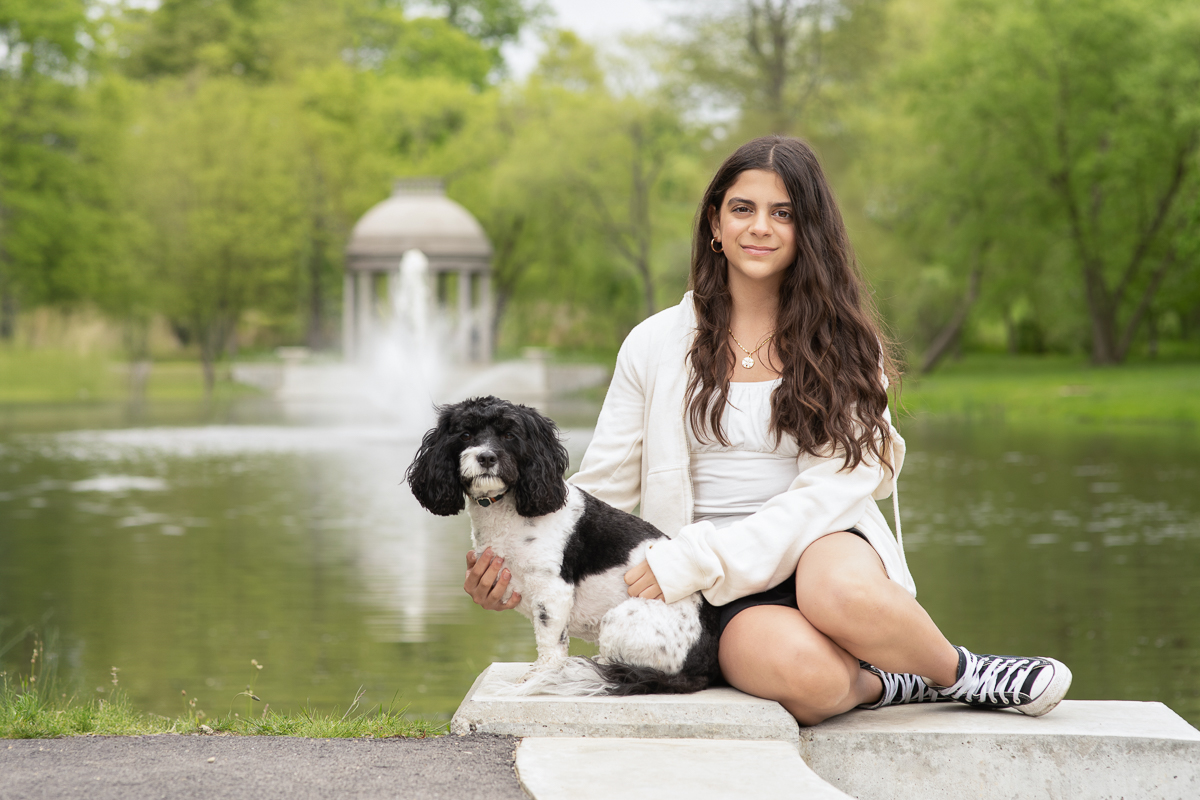 Teenage girl seated on the stone landing at the lagoon with the family dog beside her and the fountain behind at Larz Anderson Park in Brookline MA