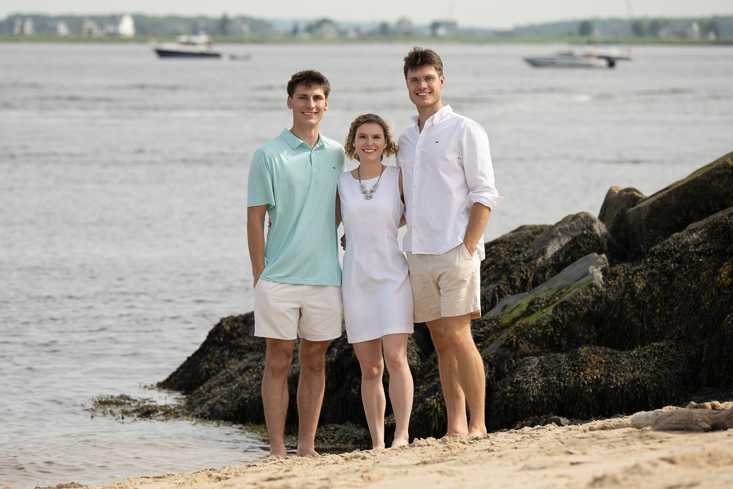 Three grown siblings standing together on the sand beside the seaweed covered rocks at the river mouth at Salisbury Beach in Salisbury Massachusetts