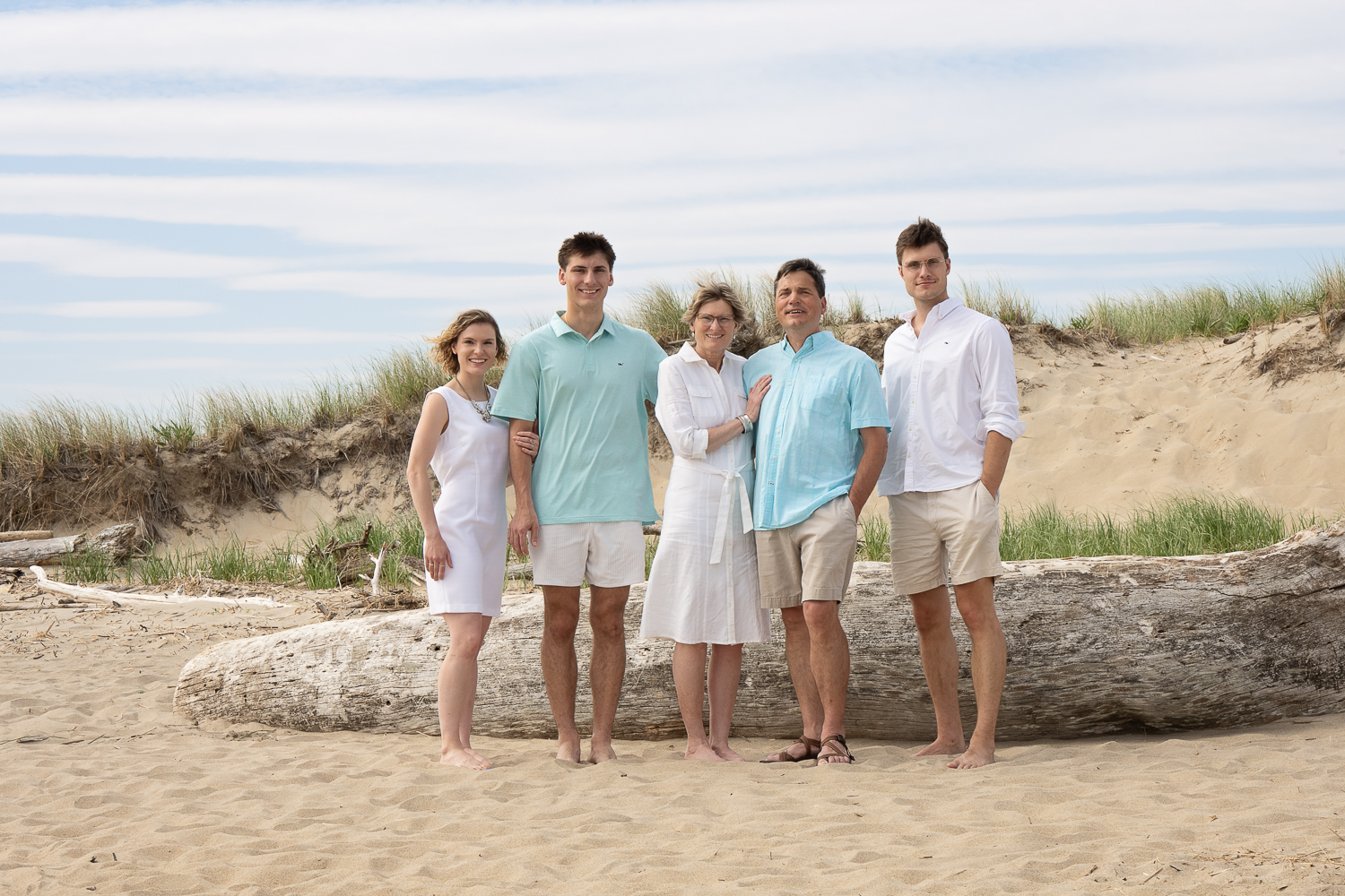 Family of five in white and pale blue standing in front of a weathered driftwood log below the dune grass at Salisbury Beach in Salisbury Massachusetts