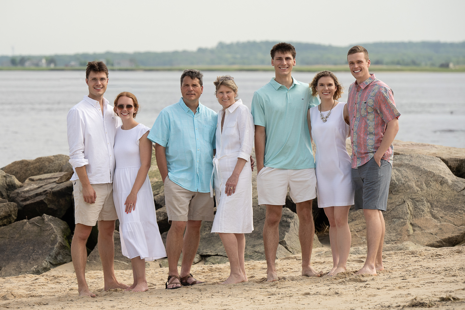 Extended family of seven barefoot on the sand with the river and the far shore behind them at Salisbury Beach State Reservation in Salisbury MA