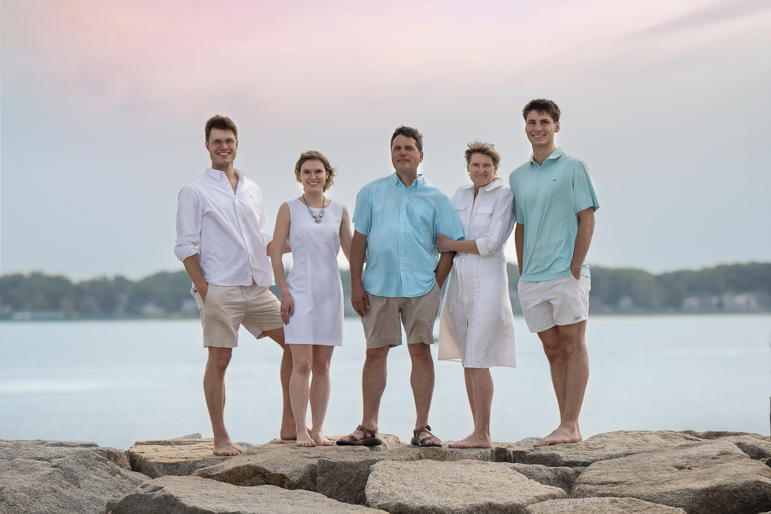 Family of five standing barefoot on the granite jetty under a pink sunset sky at Salisbury Beach State Reservation in Salisbury MA, photographed by Chelmsford family photographer Luciana Calvin