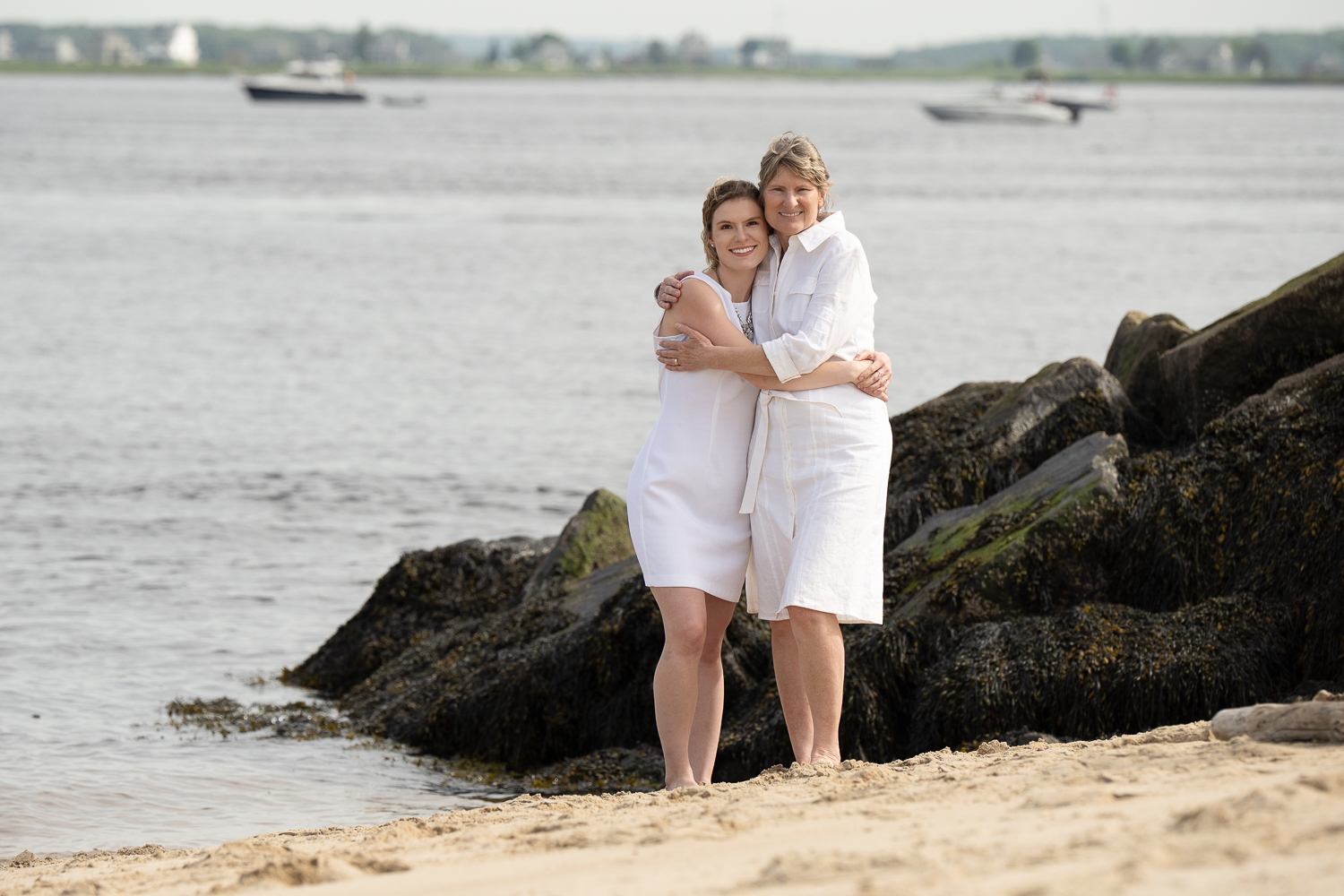 Mother and adult daughter in white embracing on the sand beside the rocks with the river behind them at Salisbury Beach State Reservation in Salisbury Massachusetts