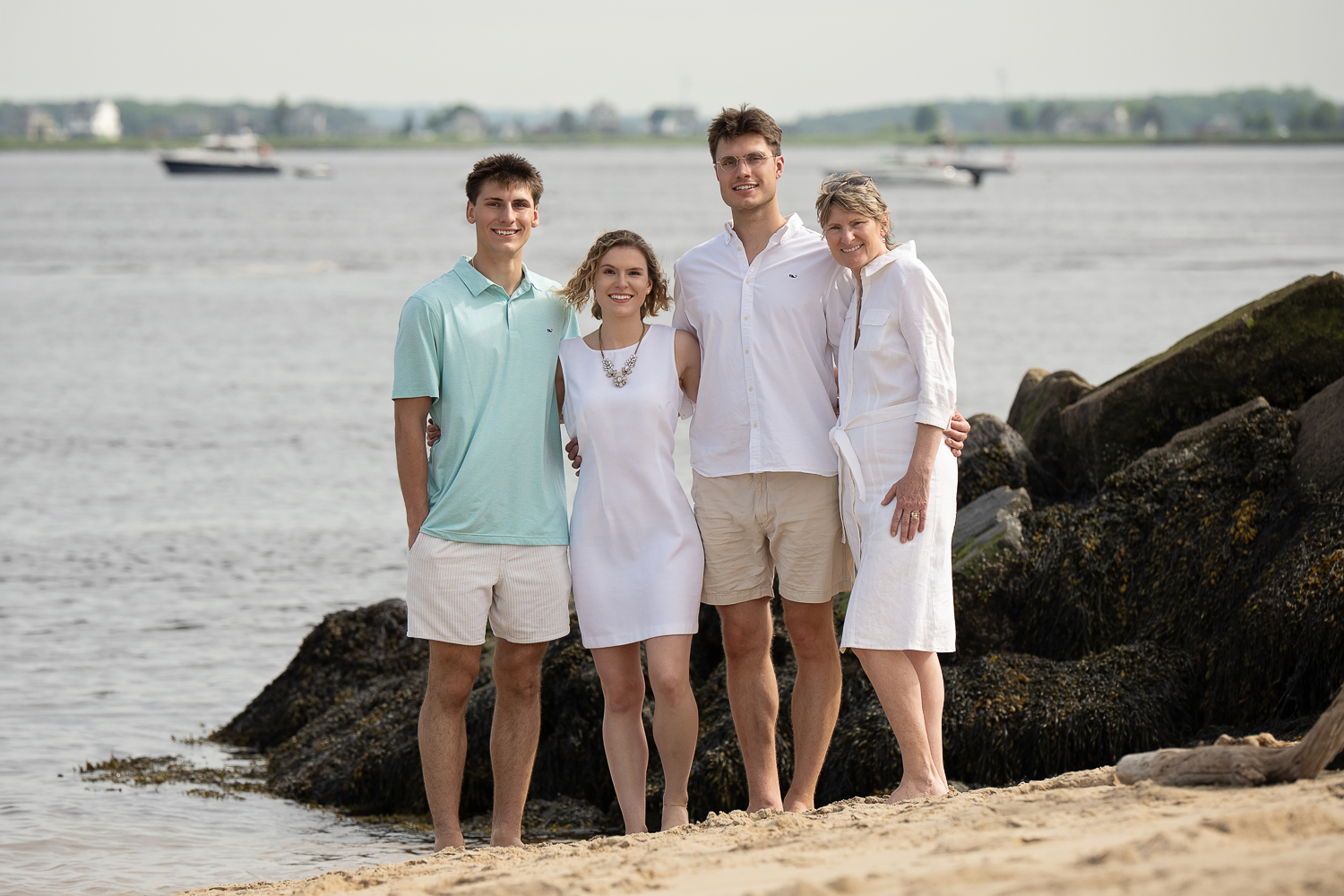 Mother with her three grown children on the rocks at the water's edge with moored boats behind them at Salisbury Beach in Salisbury MA