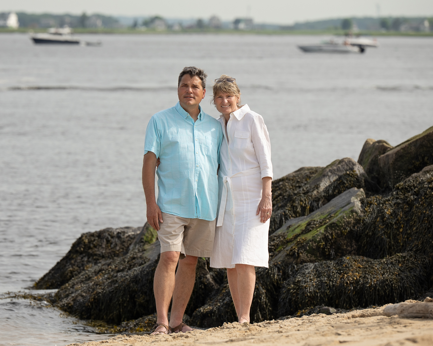 Married couple standing together on the seaweed covered rocks at the river mouth, moored boats and the far shore behind them, at Salisbury Beach State Reservation in Salisbury MA