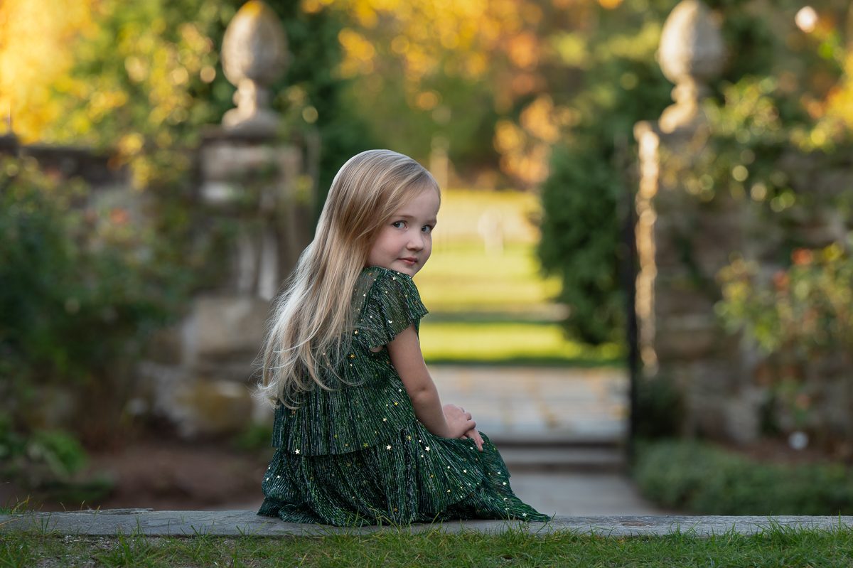 Young girl in a green dress seated on the low garden wall beside the stone piers at Stevens-Coolidge in North Andover Massachusetts