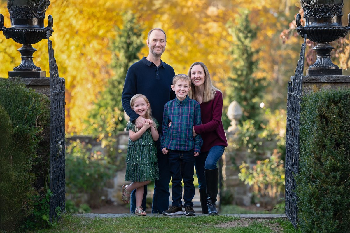 Family of four standing between the tall urn-topped stone gate piers under golden autumn trees at Stevens-Coolidge House and Gardens in North Andover MA
