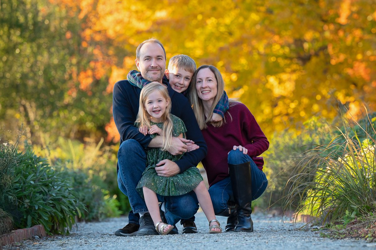 Family of four crouched together on the gravel garden path under a canopy of gold maples at Stevens-Coolidge House and Gardens in North Andover MA