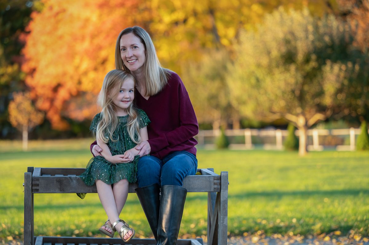 Mother seated on a garden bench with her young daughter on her lap and an orange maple behind them at Stevens-Coolidge in North Andover Massachusetts