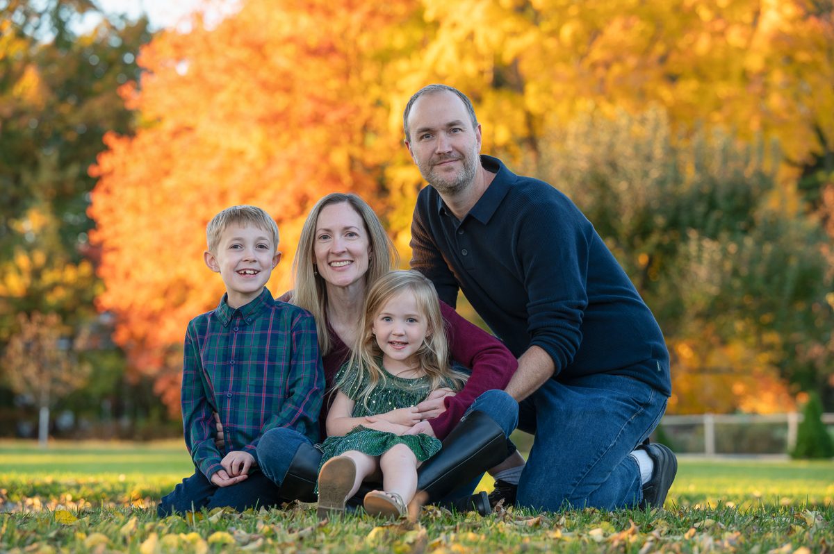 Family of four sitting together on the lawn in front of a maple in full orange at Stevens-Coolidge House and Gardens in North Andover MA