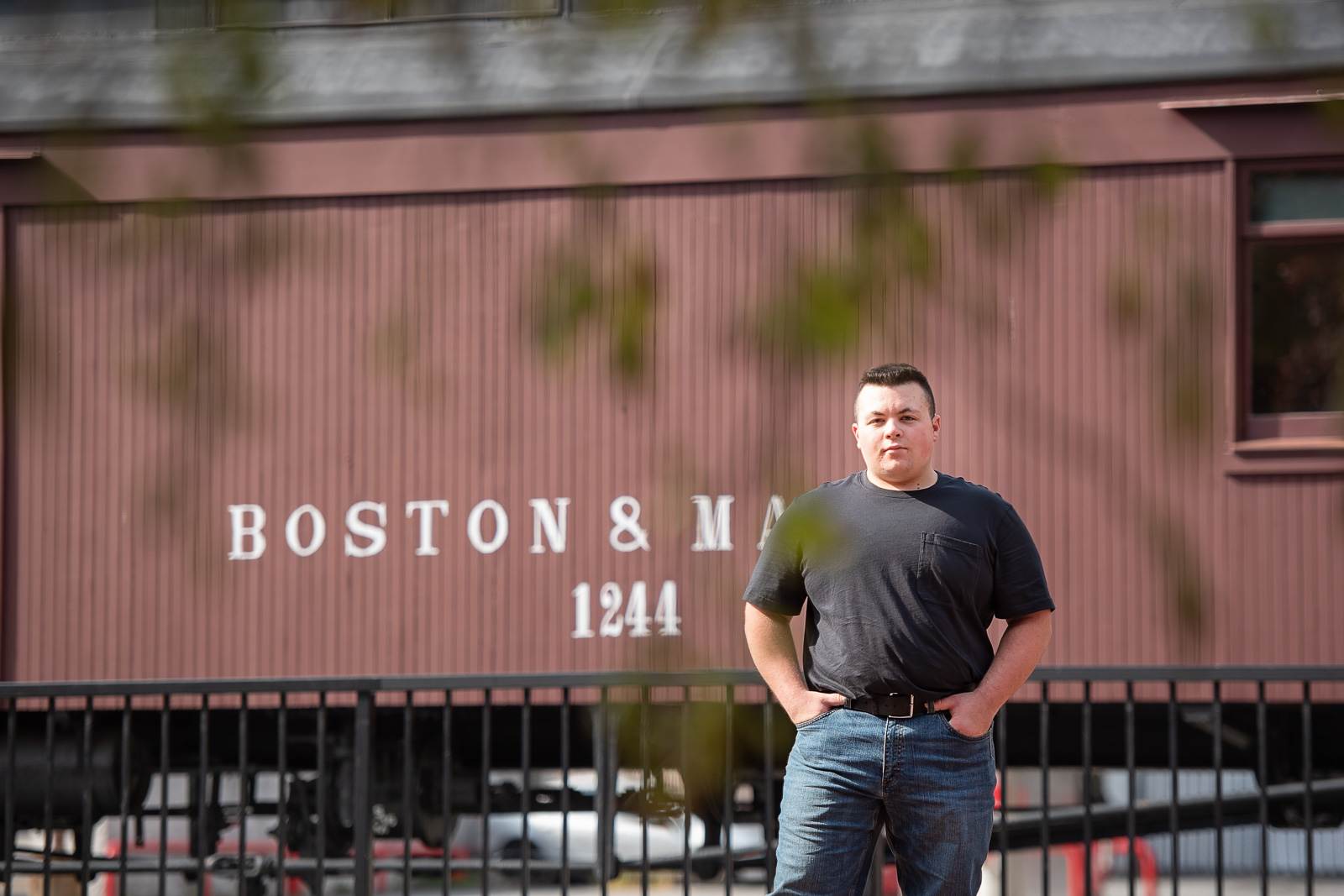 High school senior boy standing in front of a maroon Boston and Maine rail car numbered 1244 behind iron fencing in Lowell Massachusetts