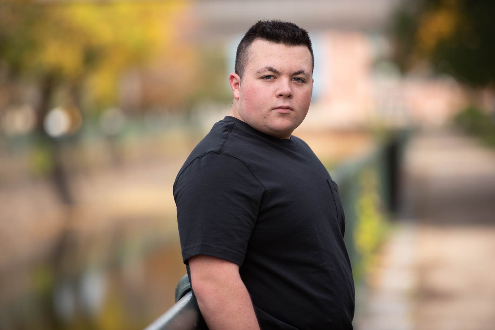 Close senior portrait of a high school boy in a black t-shirt by the canal with golden autumn color behind him in Lowell MA