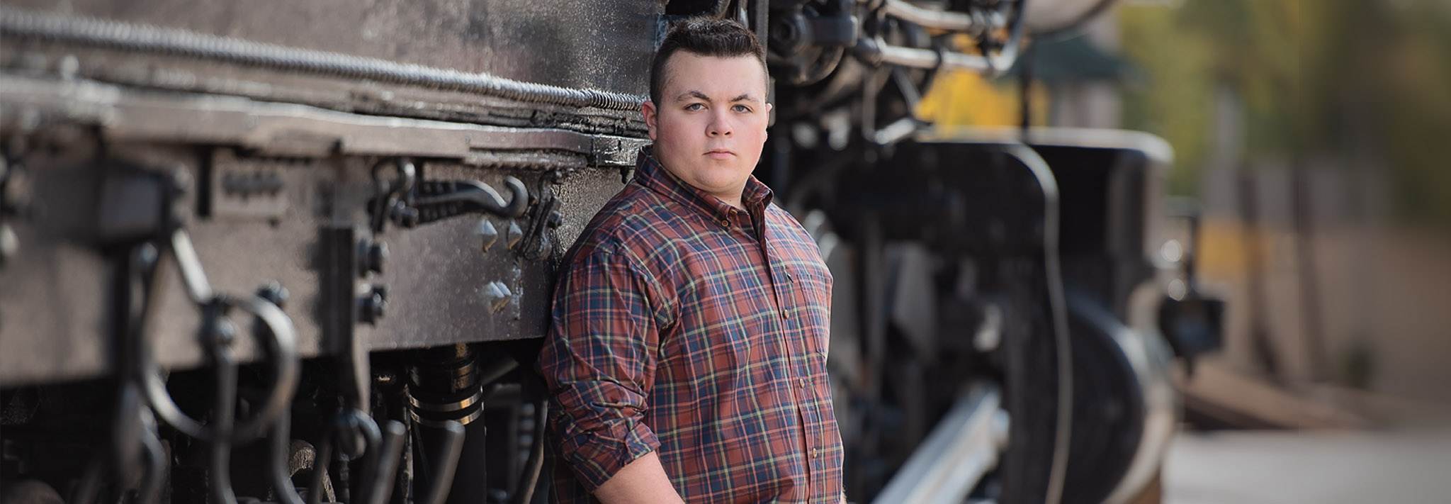 High school senior boy in a plaid button-down shirt leaning against the wheels and iron frame of a historic locomotive in Lowell MA
