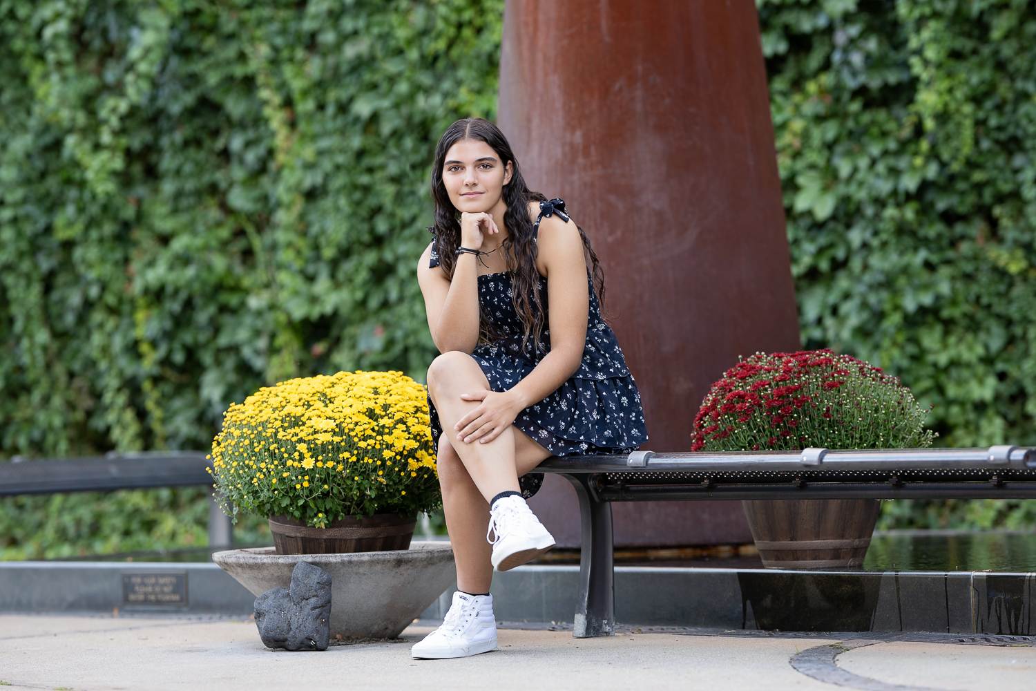 High school senior girl seated on a courtyard bench between yellow and red mums in front of an ivy wall and rust-colored sculpture in Lowell Massachusetts
