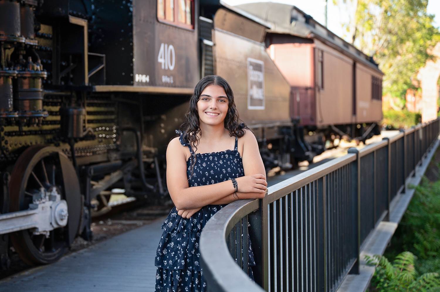 Smiling high school senior girl leaning on an iron railing beside the Boston and Maine locomotive and rail cars in Lowell Massachusetts