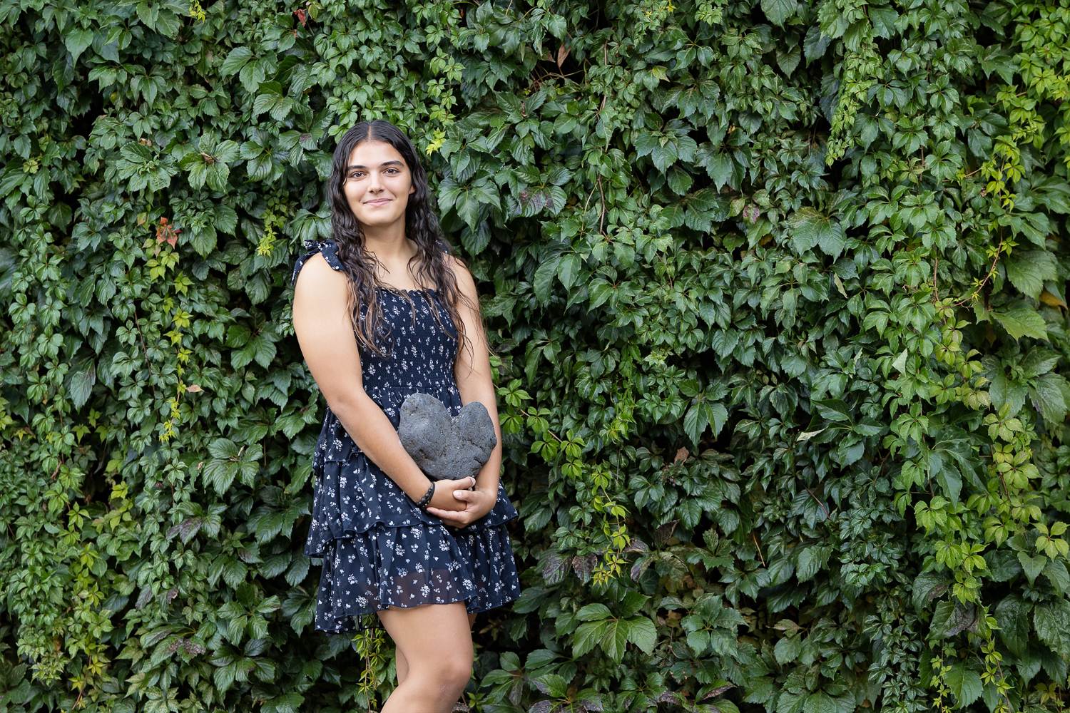 Full-length senior portrait of a high school girl in a navy dress standing in front of a tall green ivy wall in Lowell MA