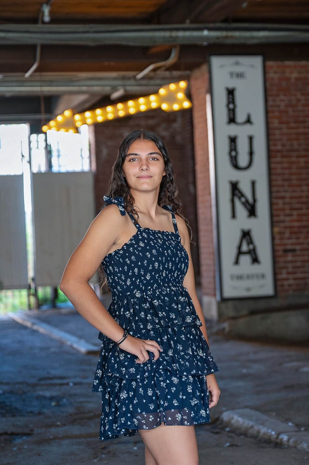 High school senior girl in a navy floral dress standing under the vertical Luna Theater sign with string lights in Lowell MA