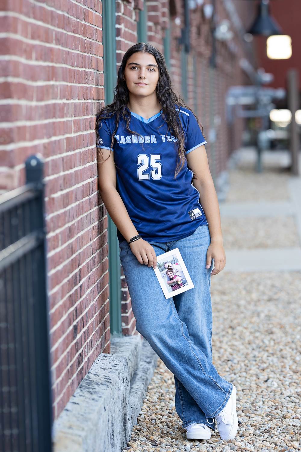 High school senior girl in her soccer jersey and jeans leaning against a brick mill building on a walkway in Lowell MA